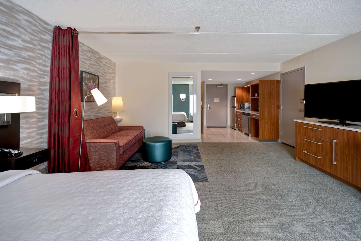 Habitación de hotel con cama, sofá, escritorio con lámpara, televisor y kitchenette.