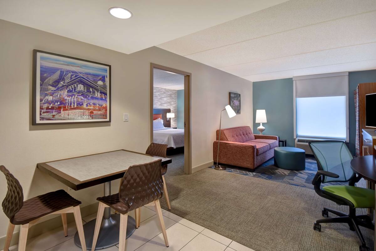 Una habitación de hotel con zona de comedor, sofá, televisor y un cuadro en la pared.