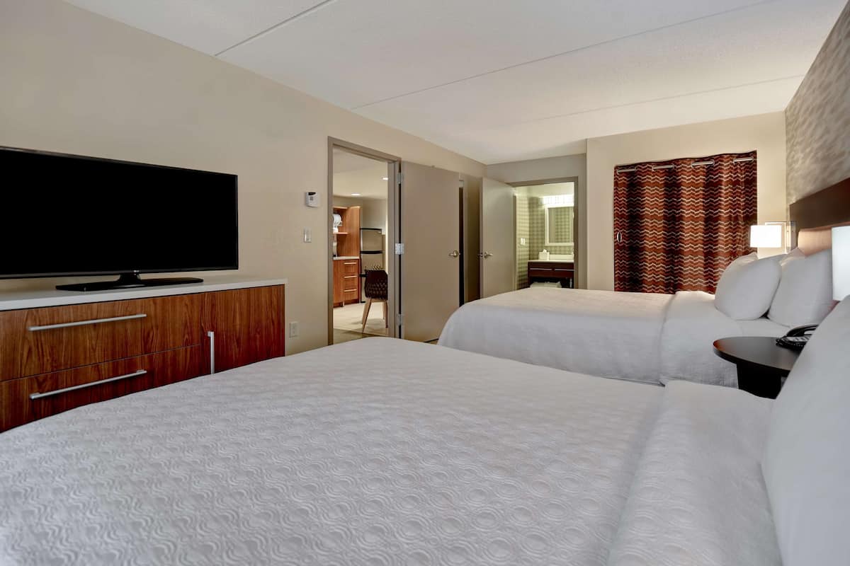 Habitación de hotel con dos camas, televisión, escritorio y baño visible a través de una puerta abierta.