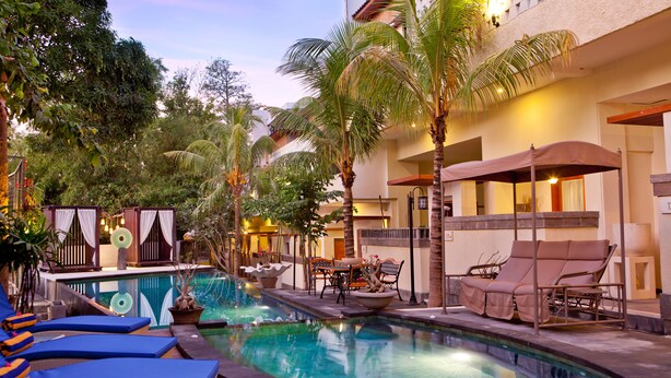 A pool area with lounge chairs and a small table, surrounded by palm trees and a building with a balcony. - Cattleya Suite by Marbella (Keroboken Kelod)