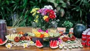 A table with a variety of fruits, vegetables, and beverages, surrounded by greenery. - Cattleya Suite by Marbella (Keroboken Kelod)
