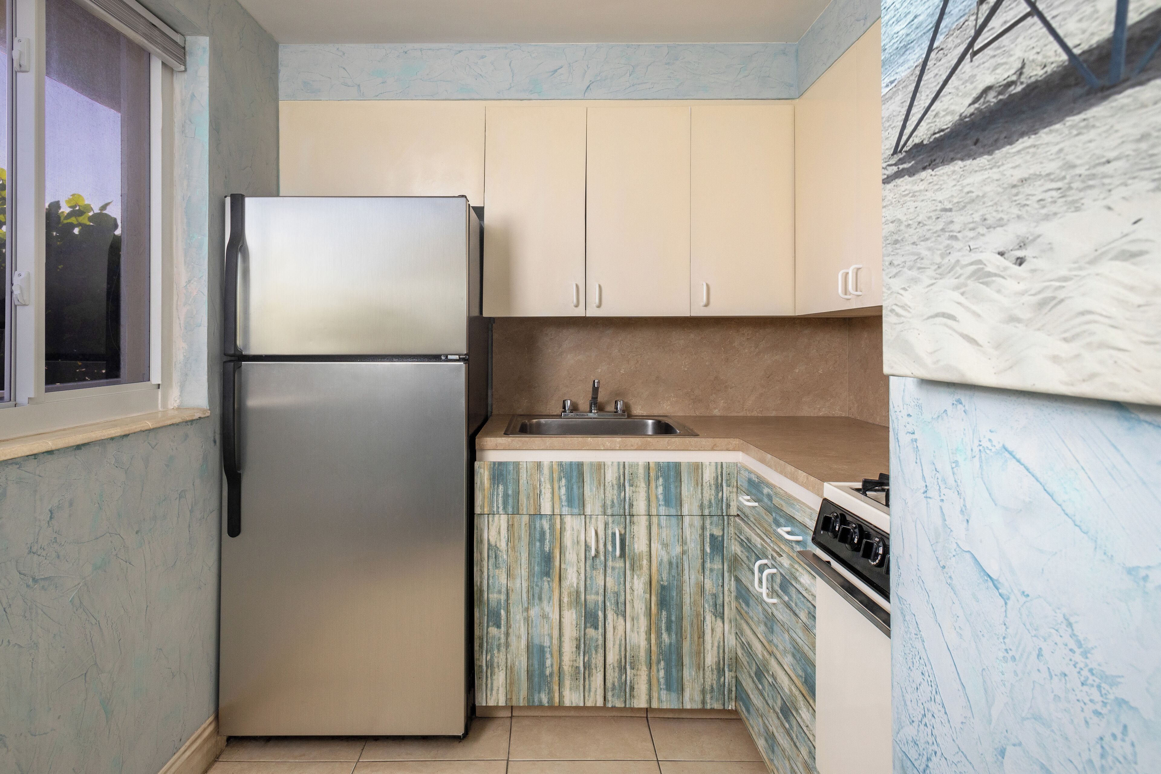 A compact kitchen with a stainless steel refrigerator, beige cabinets, a blue and white patterned backsplash, and a window with a view of greenery.