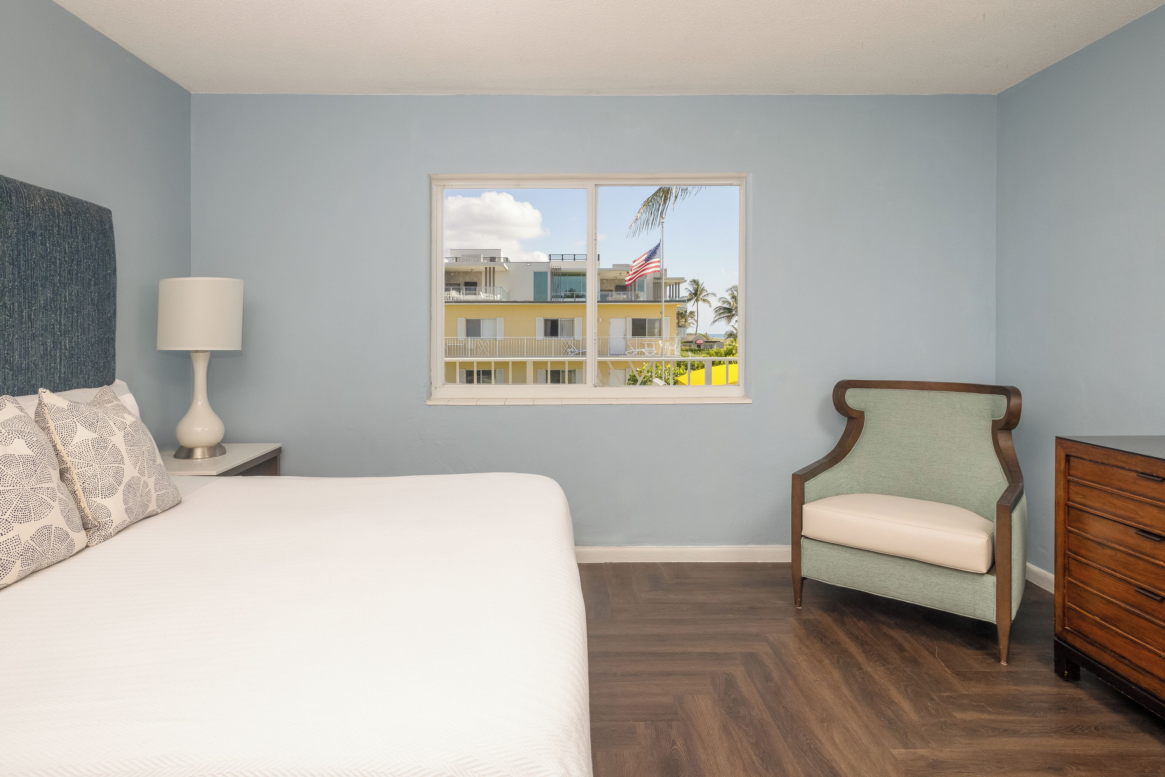 Une chambre à coucher comprenant un lit, une chaise, une commode et une fenêtre donnant sur un immeuble et des palmiers.
