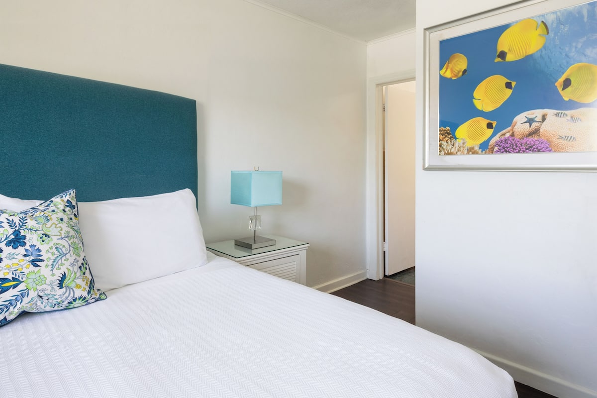 Habitación de hotel con cama, mesita de noche con lámpara, cuadro de peces en la pared y una puerta a la derecha.