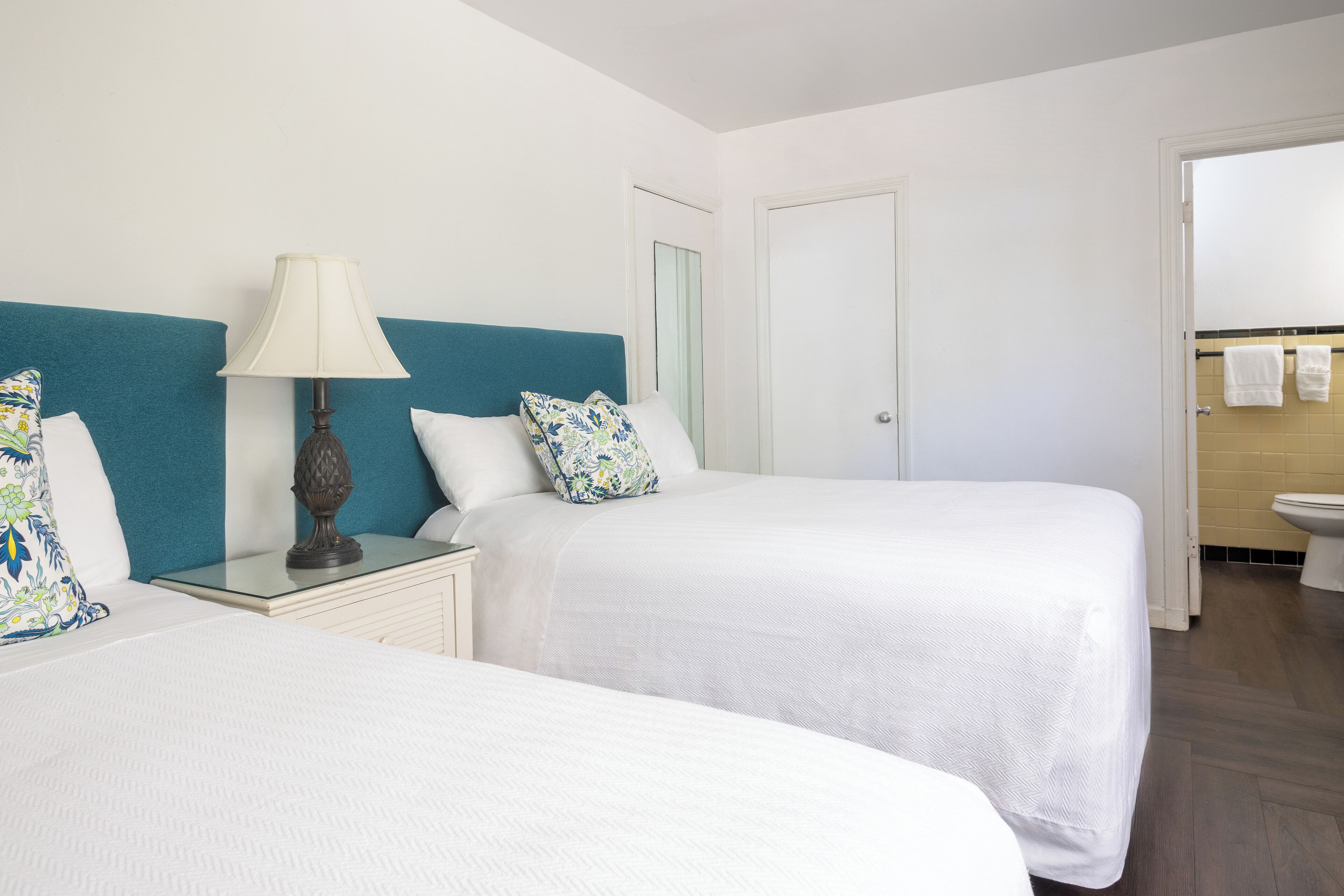 Habitación de hotel con dos camas, una mesita de noche, una lámpara y un baño con inodoro y perchero para toallas.