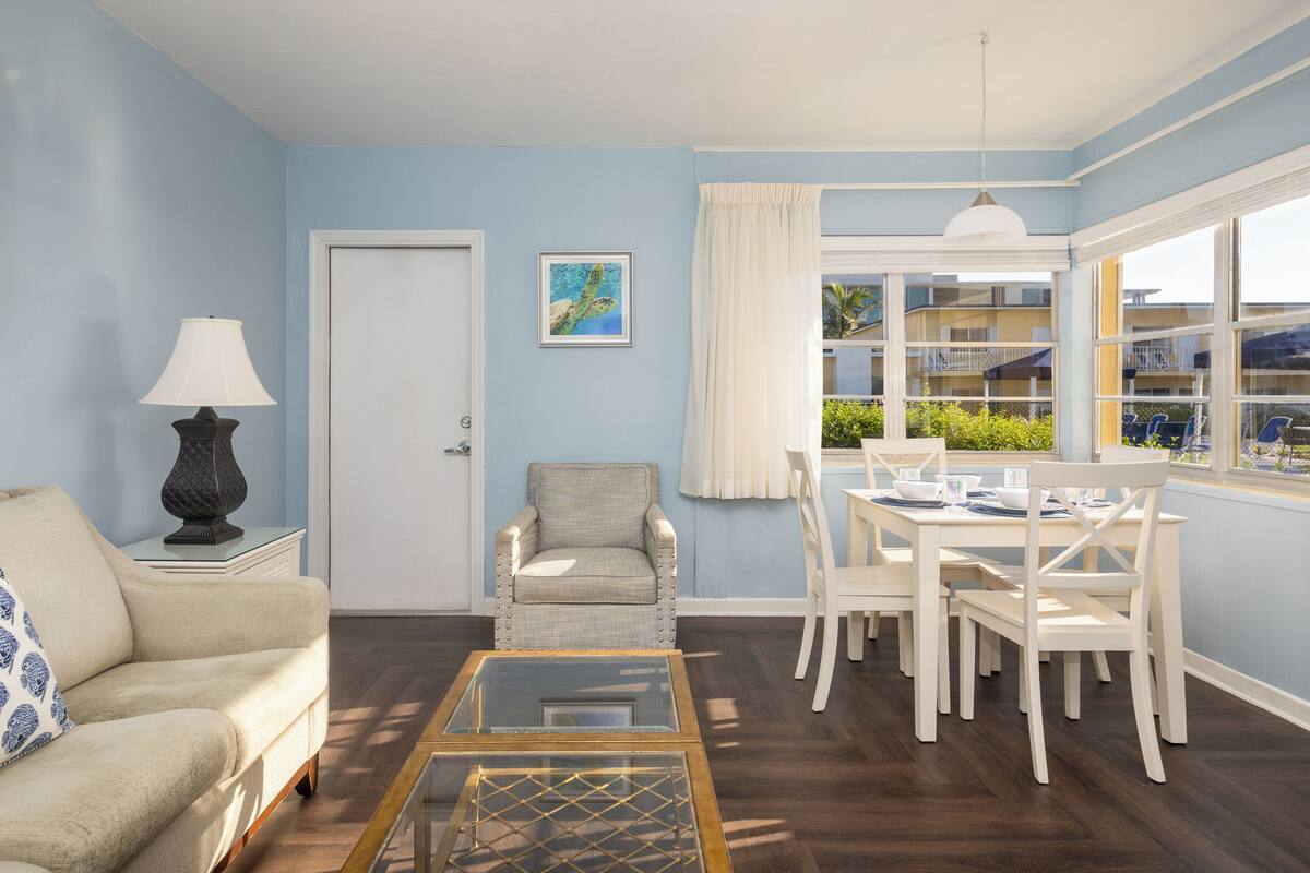 Una sala de estar con sofá, sillón, mesa de comedor con sillas, lámpara, un cuadro en la pared y ventana con cortinas.