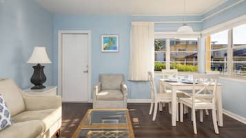 Een woonkamer met een bank, een stoel, een eettafel met stoelen, een lamp, een schilderij aan de muur en een raam met gordijnen.