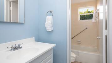 Un bagno con un lavandino bianco, uno specchio, un appendi-asciugamani, una doccia dotata di maniglione e una finestra vista verde.