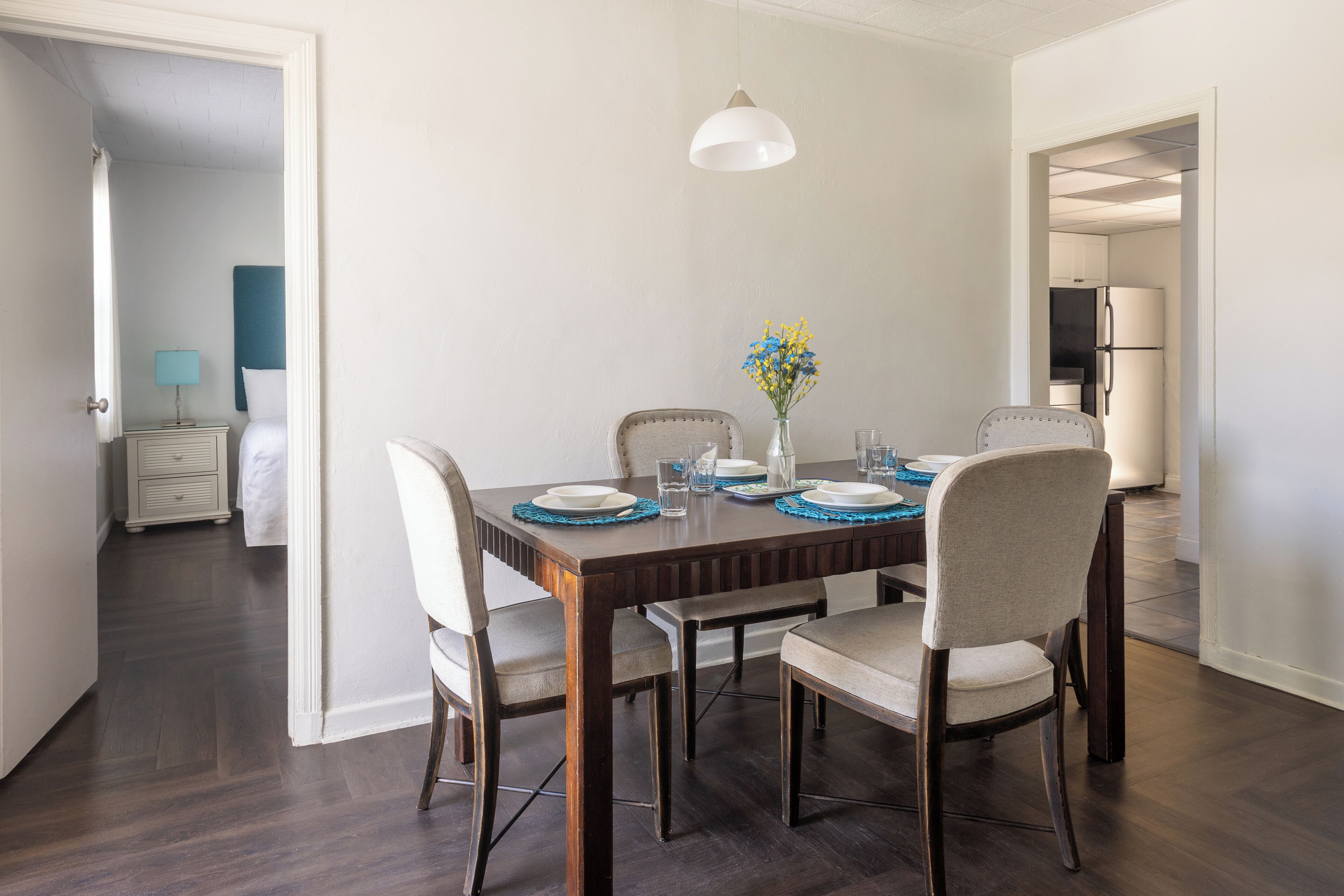 Comedor con mesa de madera dispuesta para cuatro, un florero con flores y una lámpara colgante blanca.