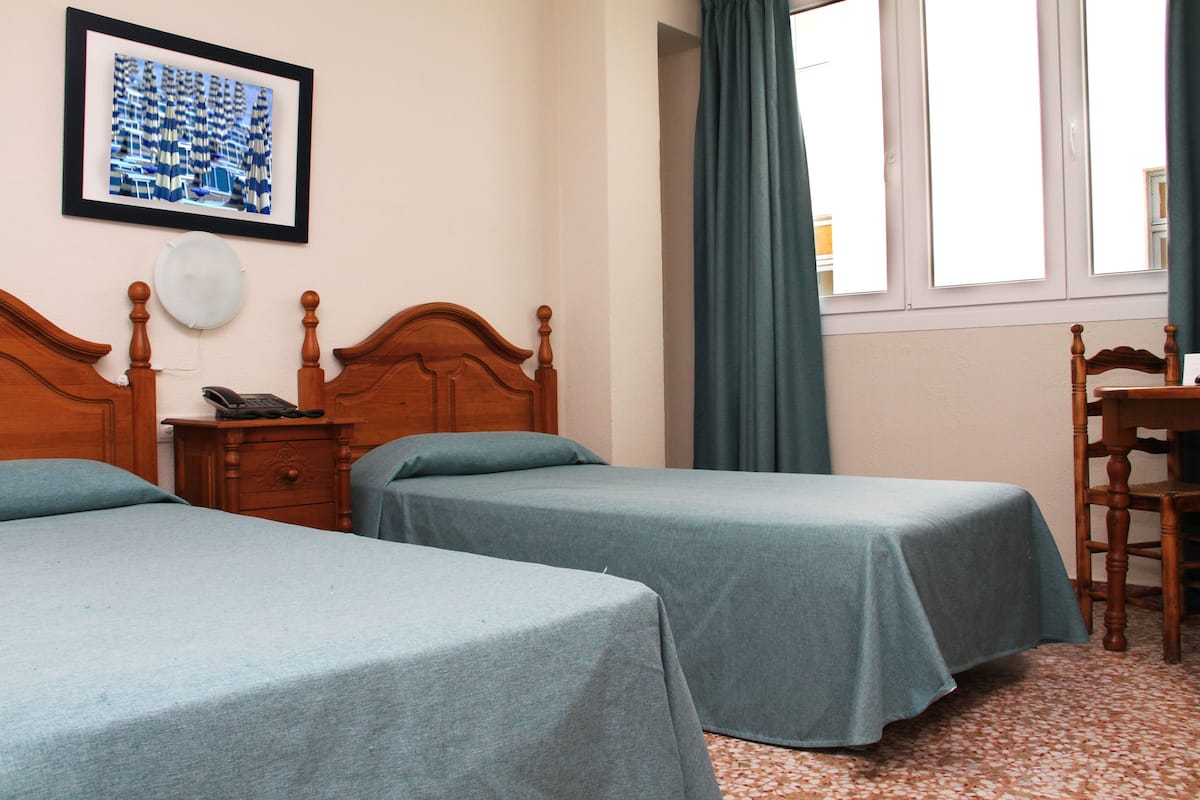 A hotel room with two single beds, a wooden nightstand with a telephone, a framed picture on the wall, and a window with curtains.