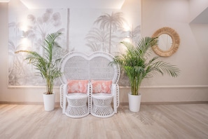 Une pièce avec une chaise en osier blanche, deux plantes en pot, une fresque murale et un miroir rond.