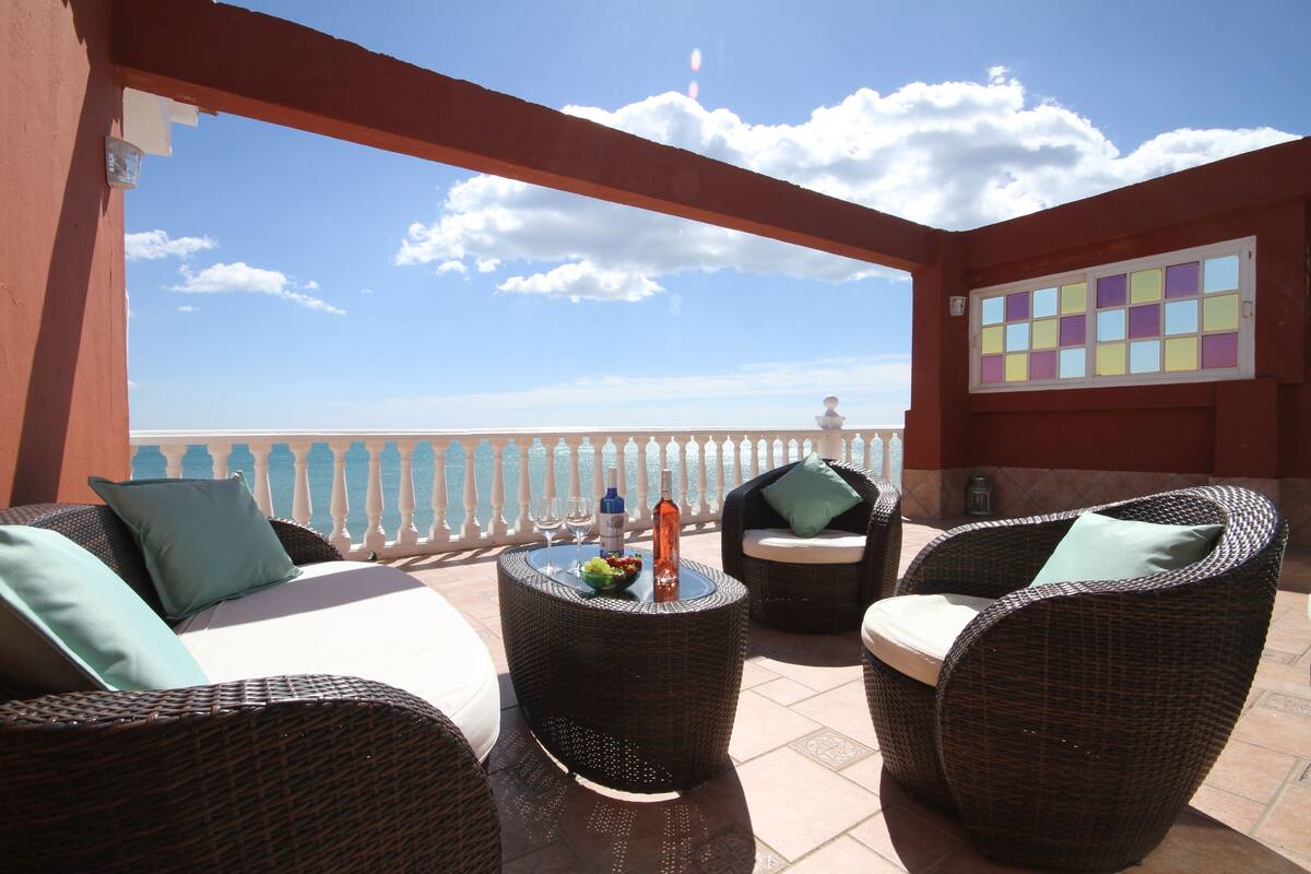 A balcony with wicker furniture, a table with a bottle and glasses, and a decorative wall.
