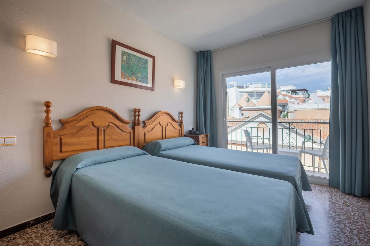 A hotel room with two beds, a large window with a view of buildings, and a balcony with chairs.