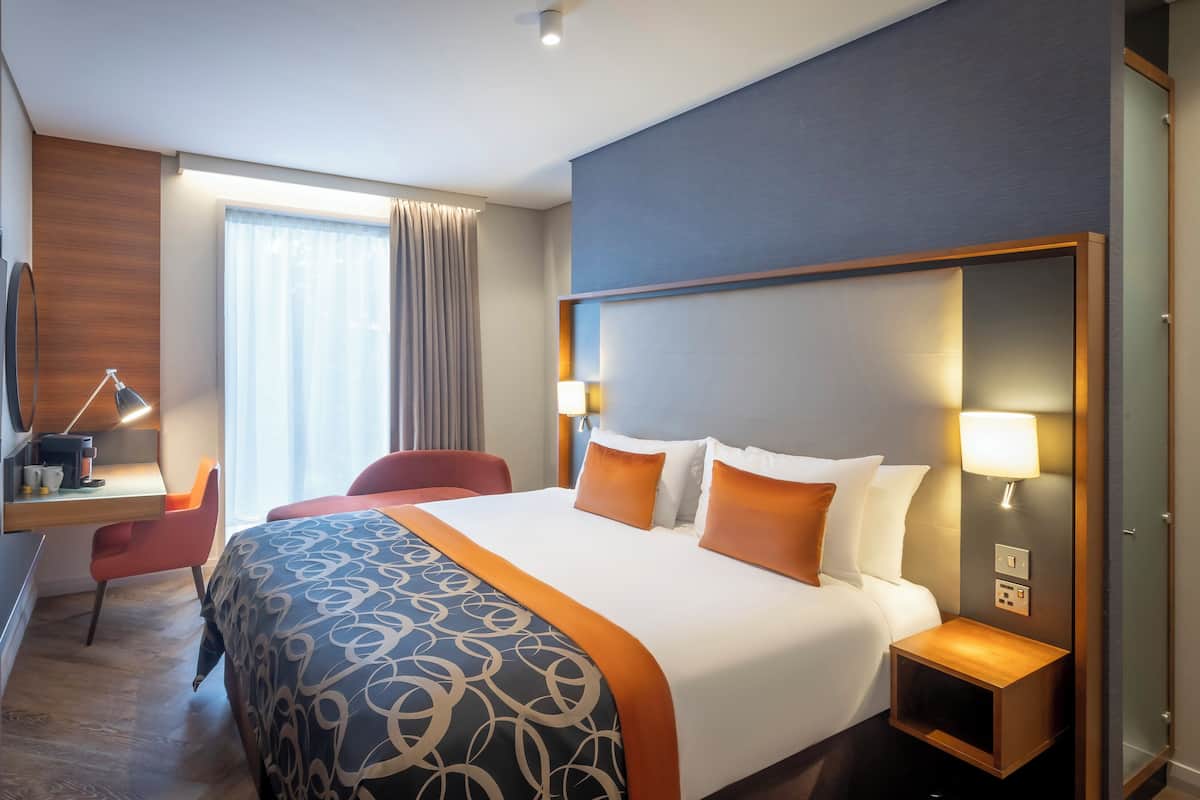 Ein Hotelzimmer mit einem Bett, Nachttischen, einem Schreibtisch mit Lampe, einem Sessel und einem Fernseher.