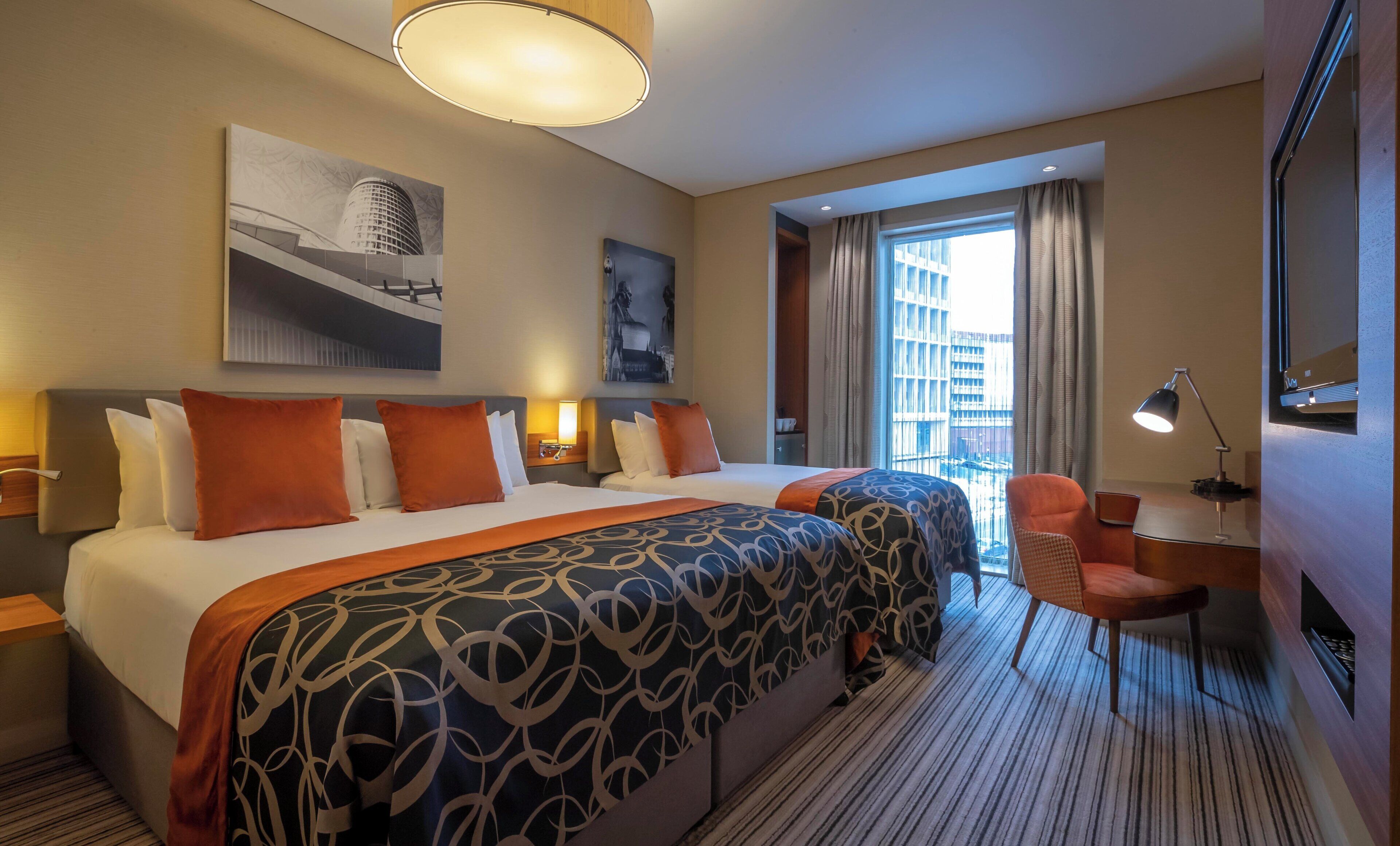 Habitación de hotel con una cama grande, un escritorio, una silla, una lámpara de escritorio y vistas a la ciudad por la ventana.