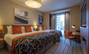 Une chambre d’hôtel avec un grand lit, un bureau, une chaise, une lampe de bureau et une vue sur la ville par la fenêtre.