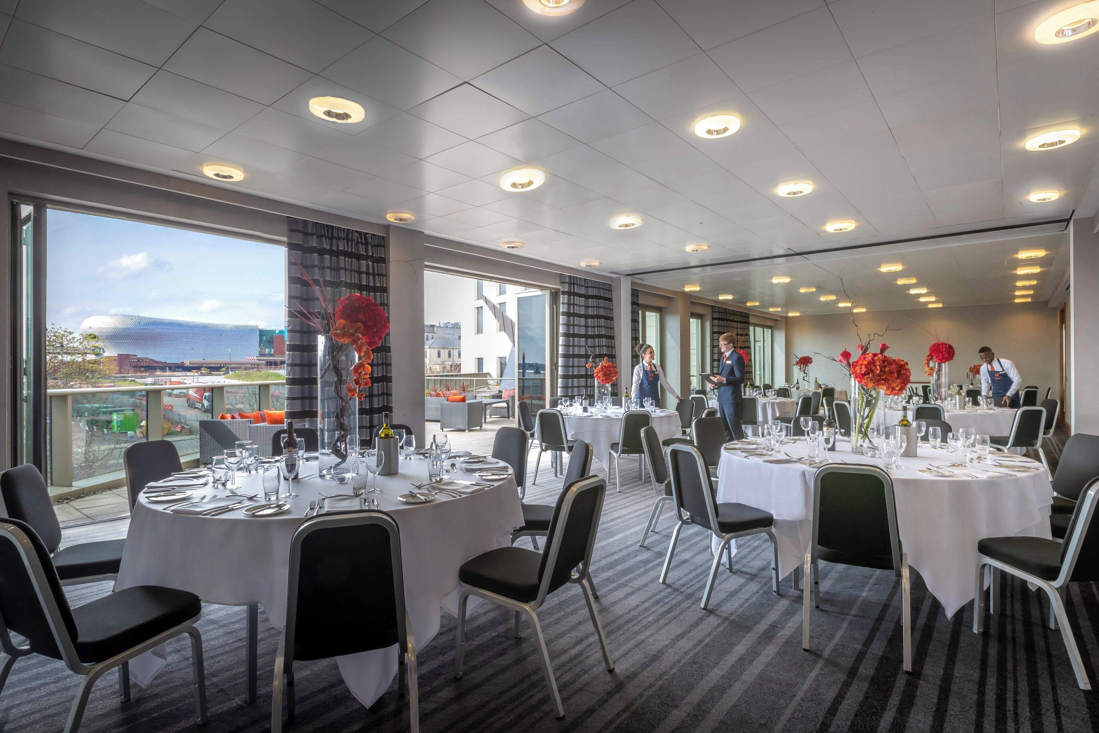 A well-lit indoor dining area with round tables, chairs, and a view of the cityscape through large windows.