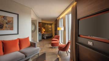 Habitación de hotel con sofá, reposapiés, televisor y vista al pasillo.