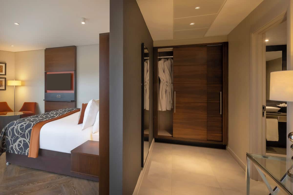 Ein modernes Hotelzimmer mit einem Bett, einer Sitzecke mit Stuhl und Tisch, einem Kleiderschrank und einem Badezimmer mit Glastrennwand.
