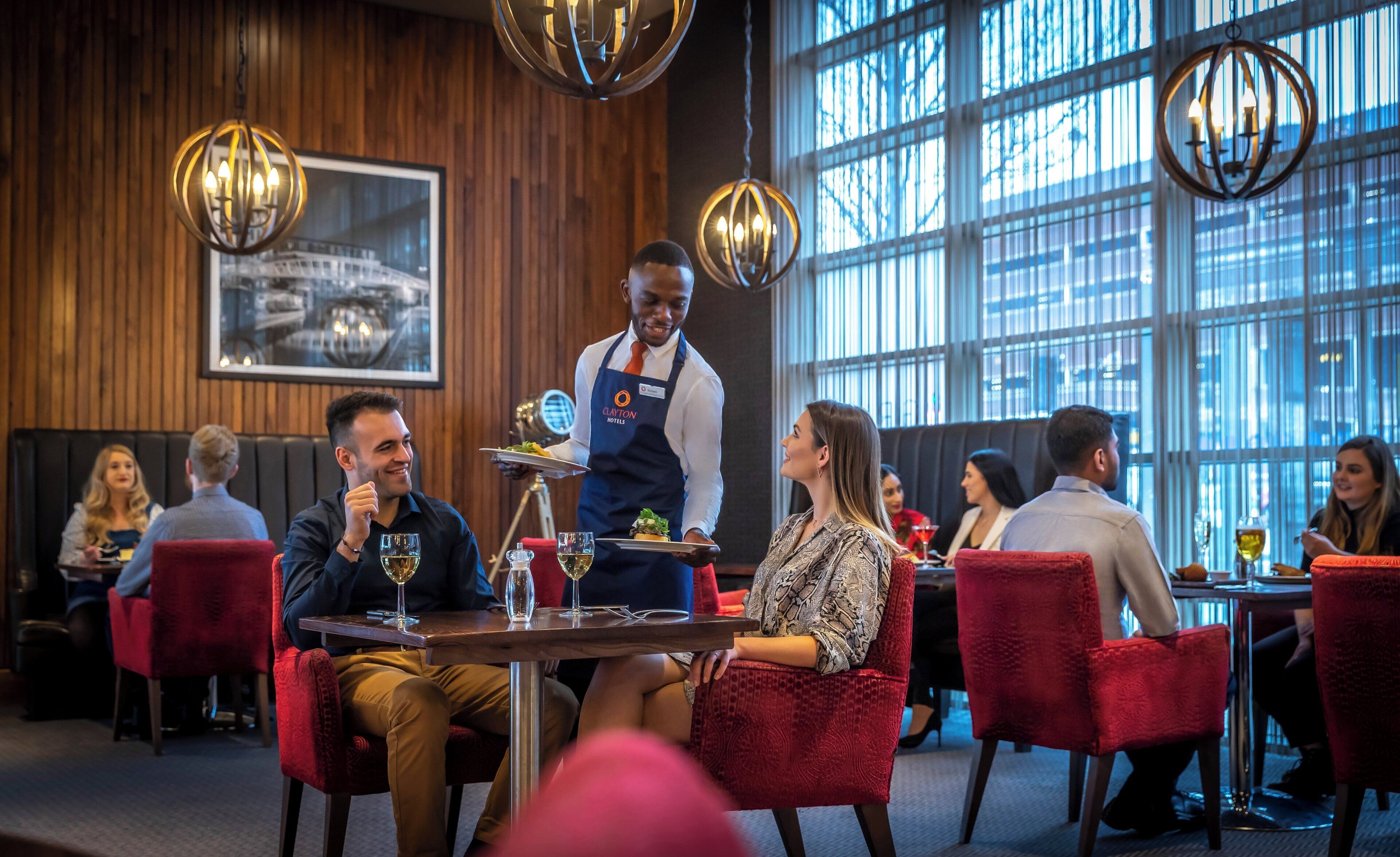 A restaurant interior with wooden paneling, red upholstered chairs, and a waiter serving a table.