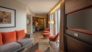 Ein modernes Hotelzimmer mit einem grauen Sofa, orangefarbenen Kissen, einem Fernseher und einem Essbereich im Hintergrund.