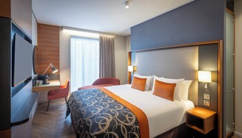 A hotel room with a bed, bedside lamps, a desk with a lamp, a chair, and a television.