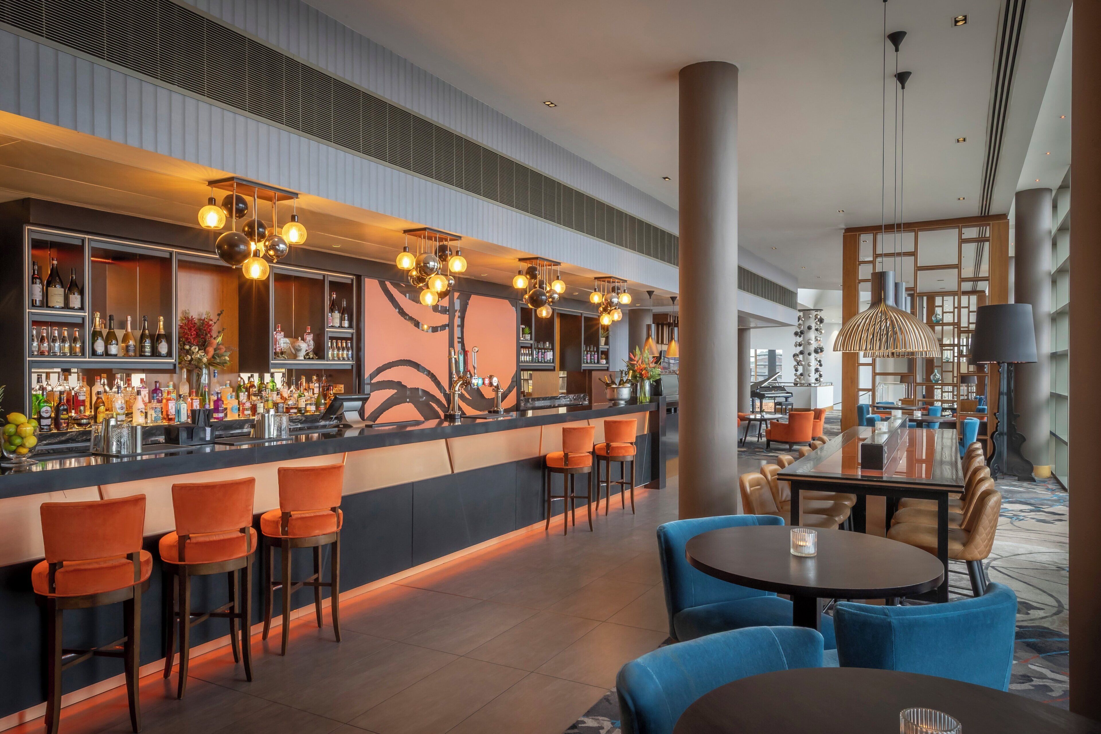 Zona de bar con diversas opciones de asiento, incluyendo taburetes y mesas redondas, y una variedad de botellas y copas en las estanterías.