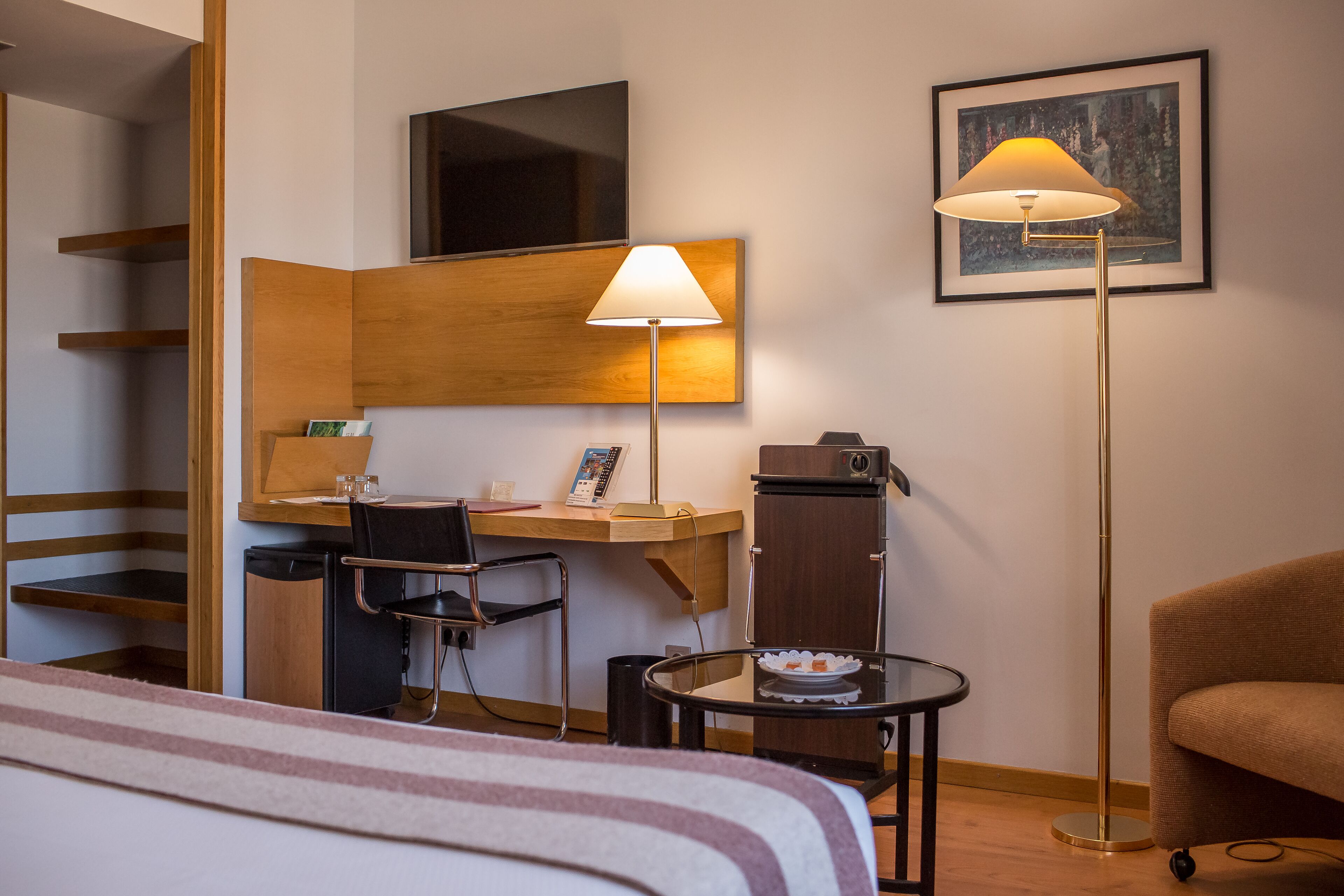 A hotel room with a bed, a desk with a chair, a small table with a plate, a floor lamp, a TV, and a framed picture on the wall.