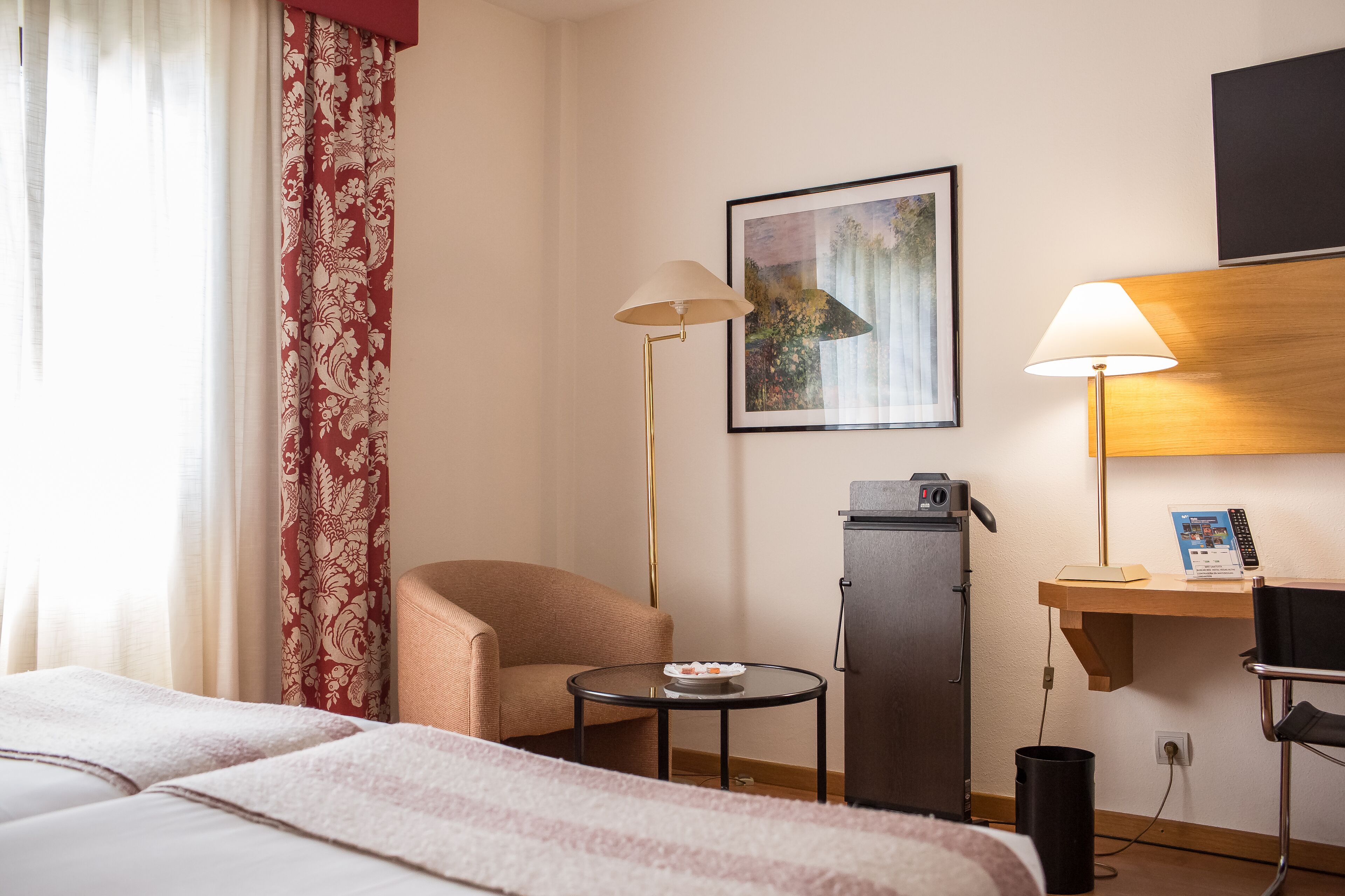 Een hotelkamer met een bed, een stoel, een klein tafeltje met een fruitschaal, een bureau met een lamp, een koelkast en een foto aan de muur.
