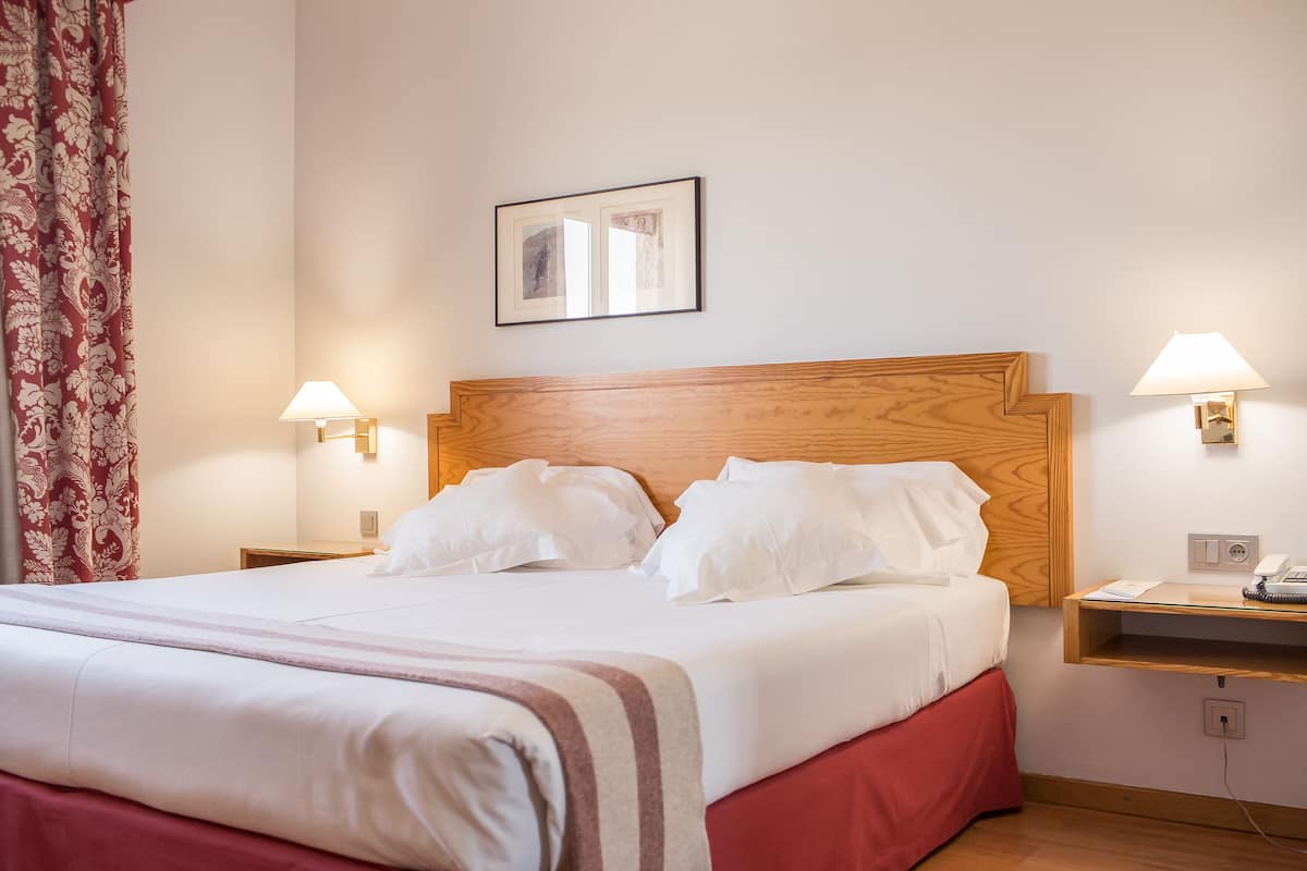 A neatly made bed with white linens and a red skirt, two bedside lamps, a wooden headboard, and a framed picture on the wall.