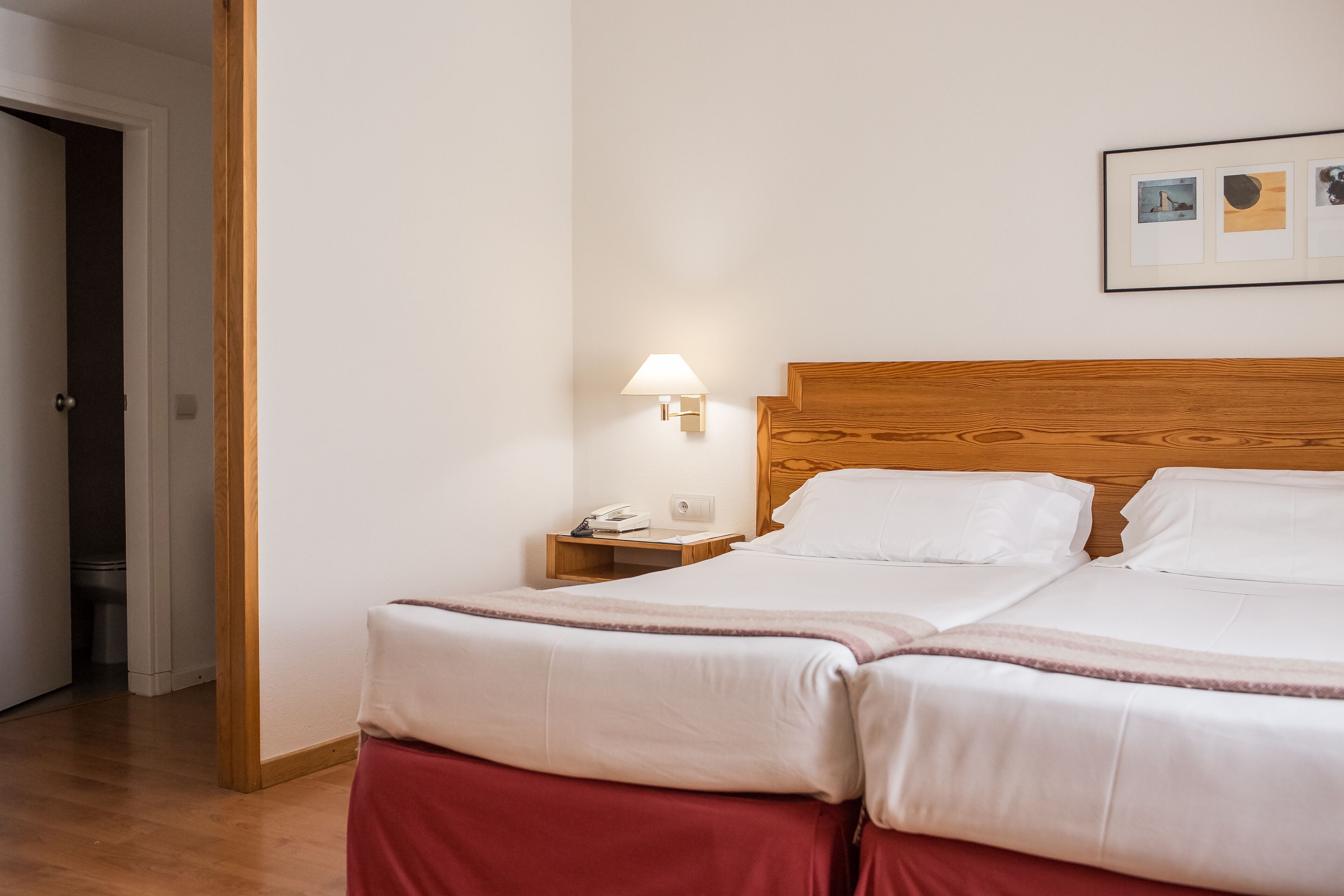 A double bed with white linens, a wooden headboard, a nightstand with a lamp, and a framed picture on the wall.
