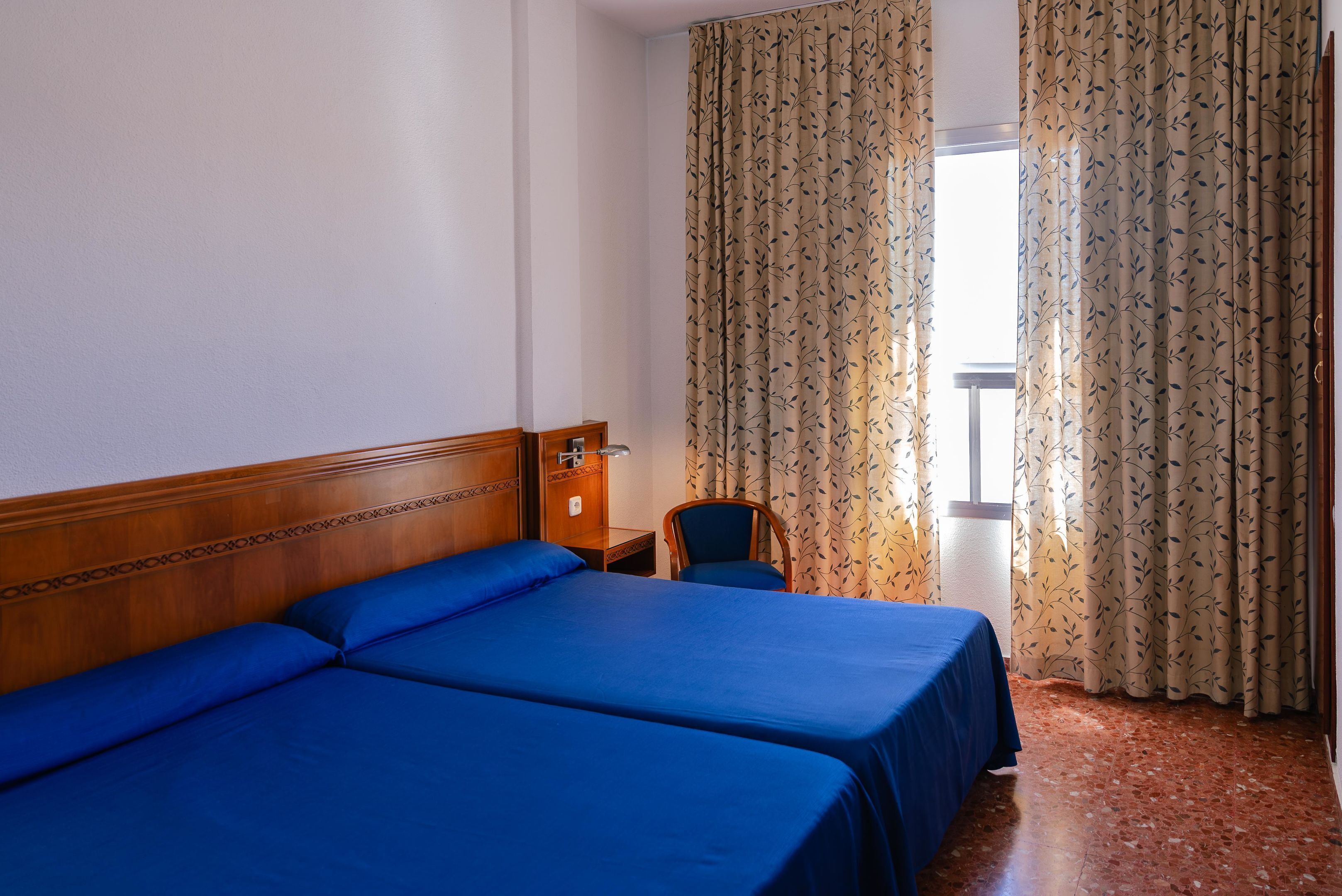 A hotel room with two beds, a wooden headboard, a small table with a lamp, a chair, and a window with curtains.