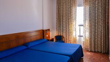 A hotel room with two beds, a wooden headboard, a small table with a lamp, a chair, and a window with curtains.