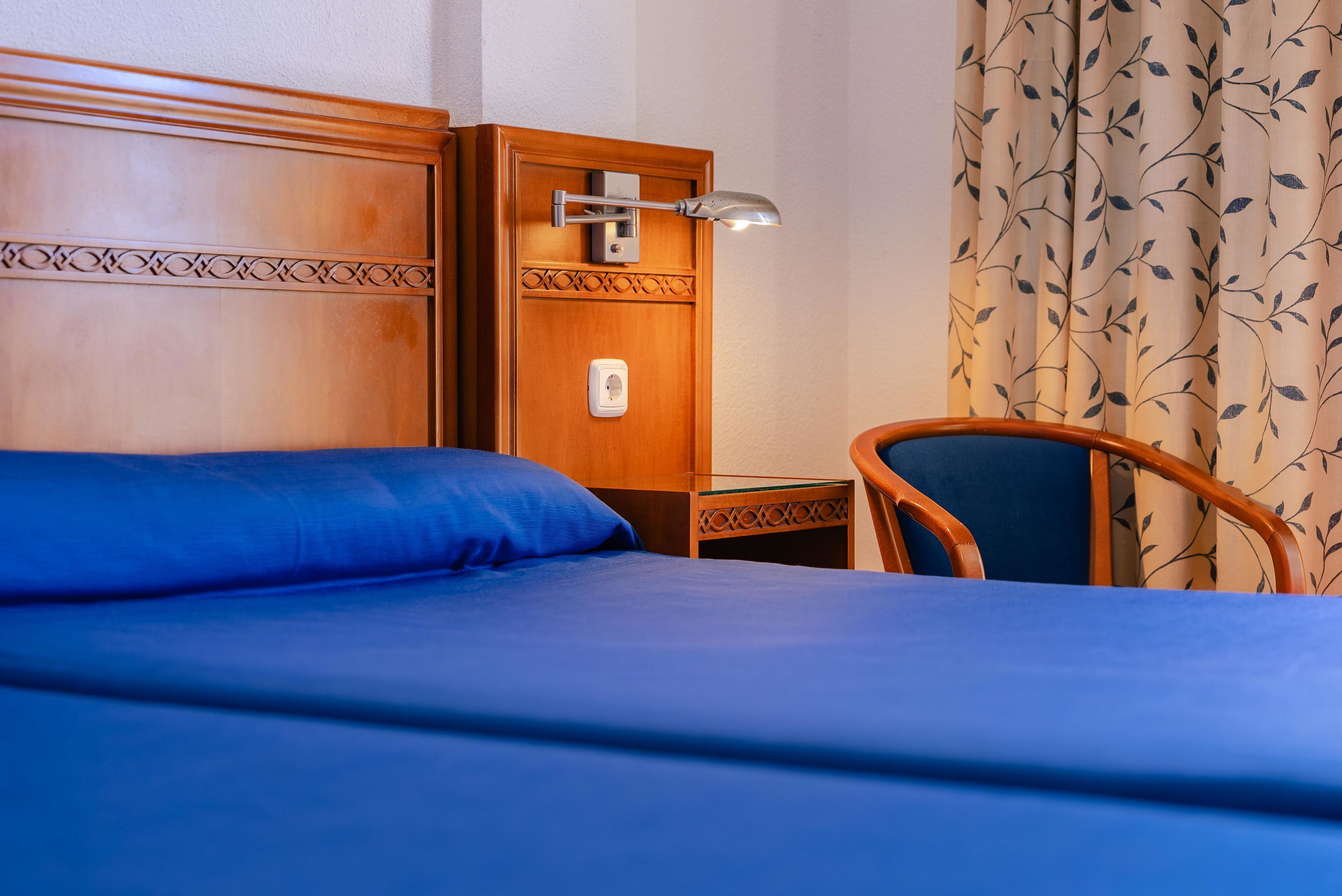 A hotel room with a wooden headboard, a single bed with blue bedding, a nightstand with a lamp, and a chair.