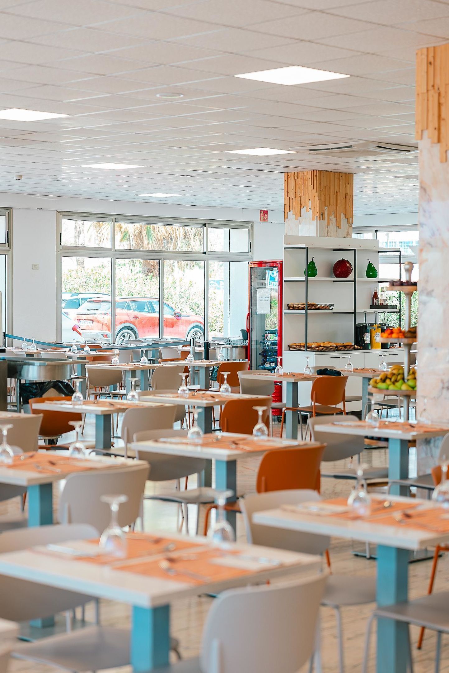 A spacious dining area with multiple tables and chairs, large windows with a view of parked cars, and a kitchen area visible in the background.