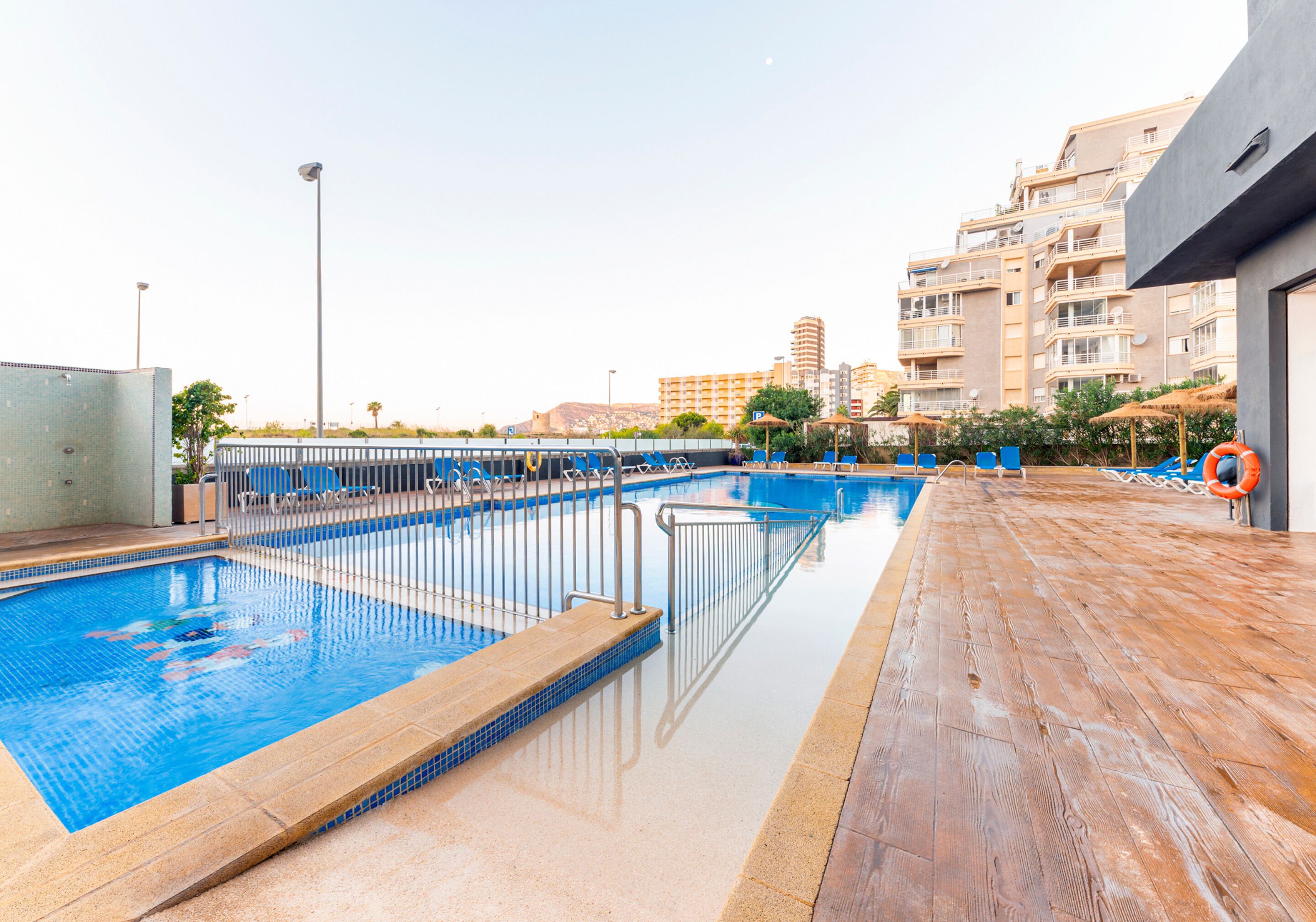 Una piscina al aire libre con muelle de madera, rodeada de sillas de descanso y sombrillas, con edificios al fondo.
