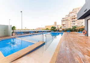 An outdoor swimming pool with a wooden deck, surrounded by lounge chairs and umbrellas, with buildings in the background.