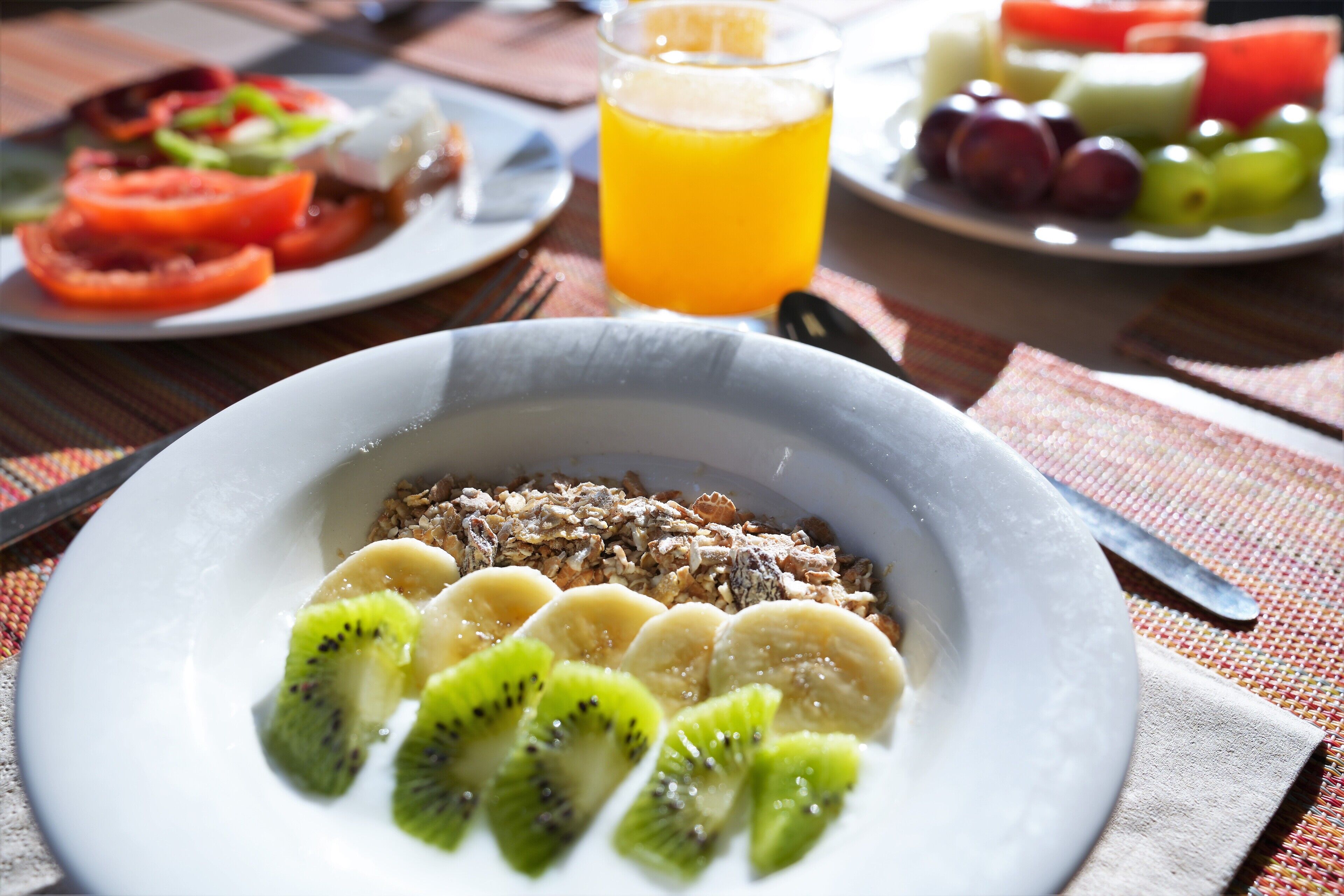 Un desayuno con un plato de cereal, rodajas de plátano y kiwi, un vaso de jugo de naranja, y un plato con diversas frutas.