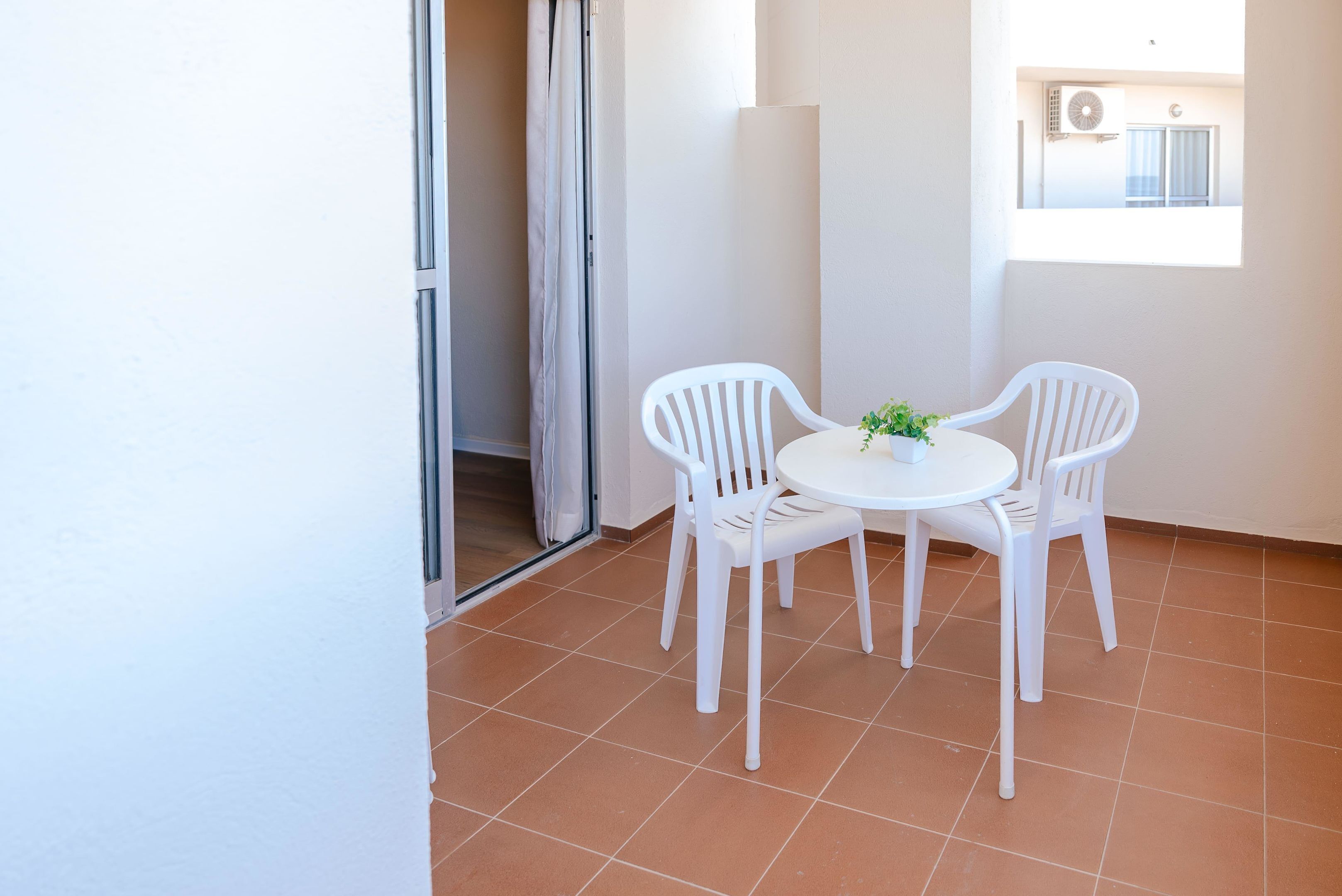 Un pequeño balcón con sillas y una mesa blancos, una puerta corrediza de vidrio y vistas a un edificio con un equipo de aire acondicionado.
