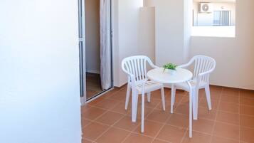 Un pequeño balcón con sillas y una mesa blancos, una puerta corrediza de vidrio y vistas a un edificio con un equipo de aire acondicionado.