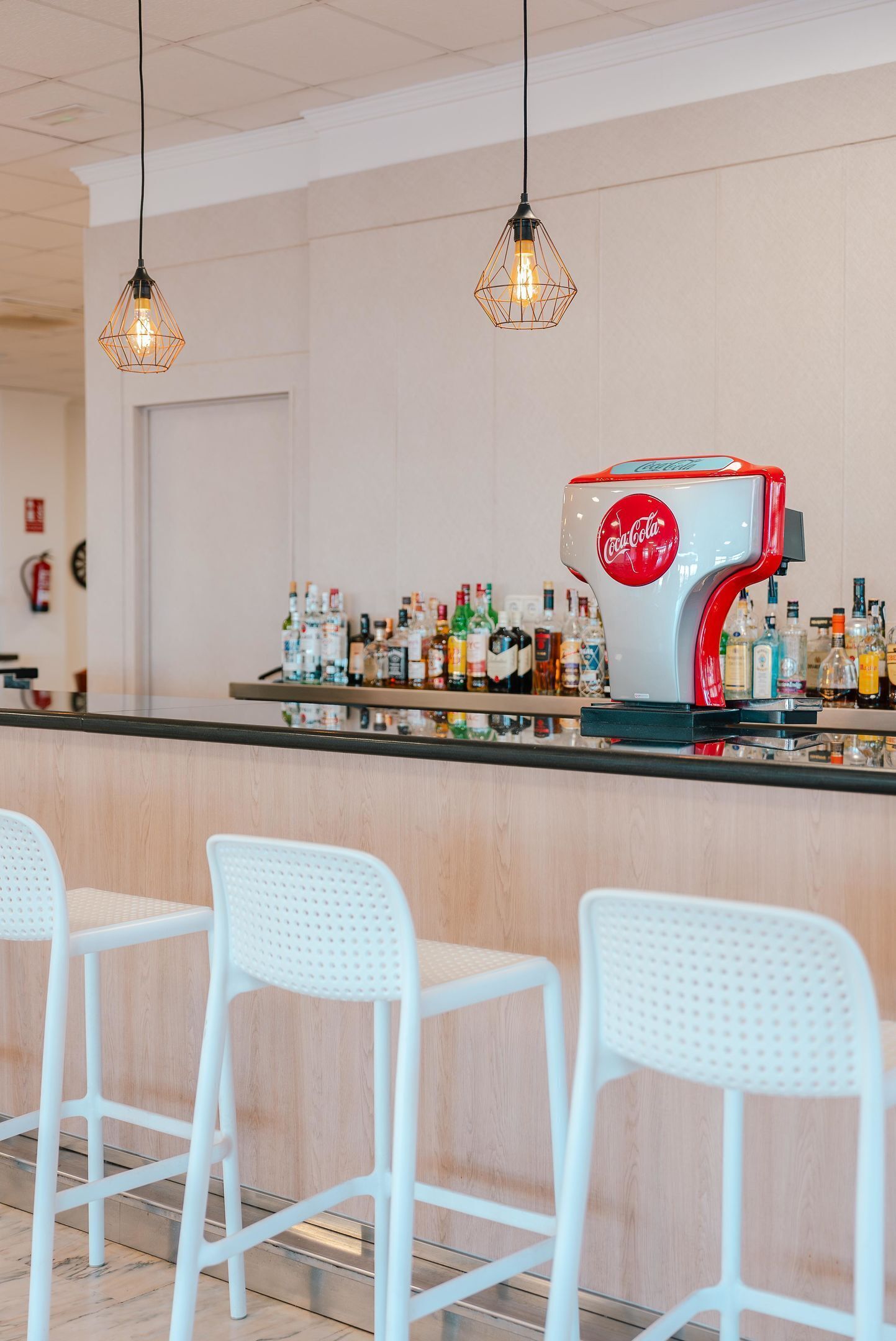 A bar area with a Coca-Cola branded coffee machine, white bar stools, and hanging pendant lights.