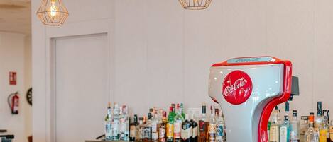 A bar area with a Coca-Cola branded coffee machine, white bar stools, and hanging pendant lights.