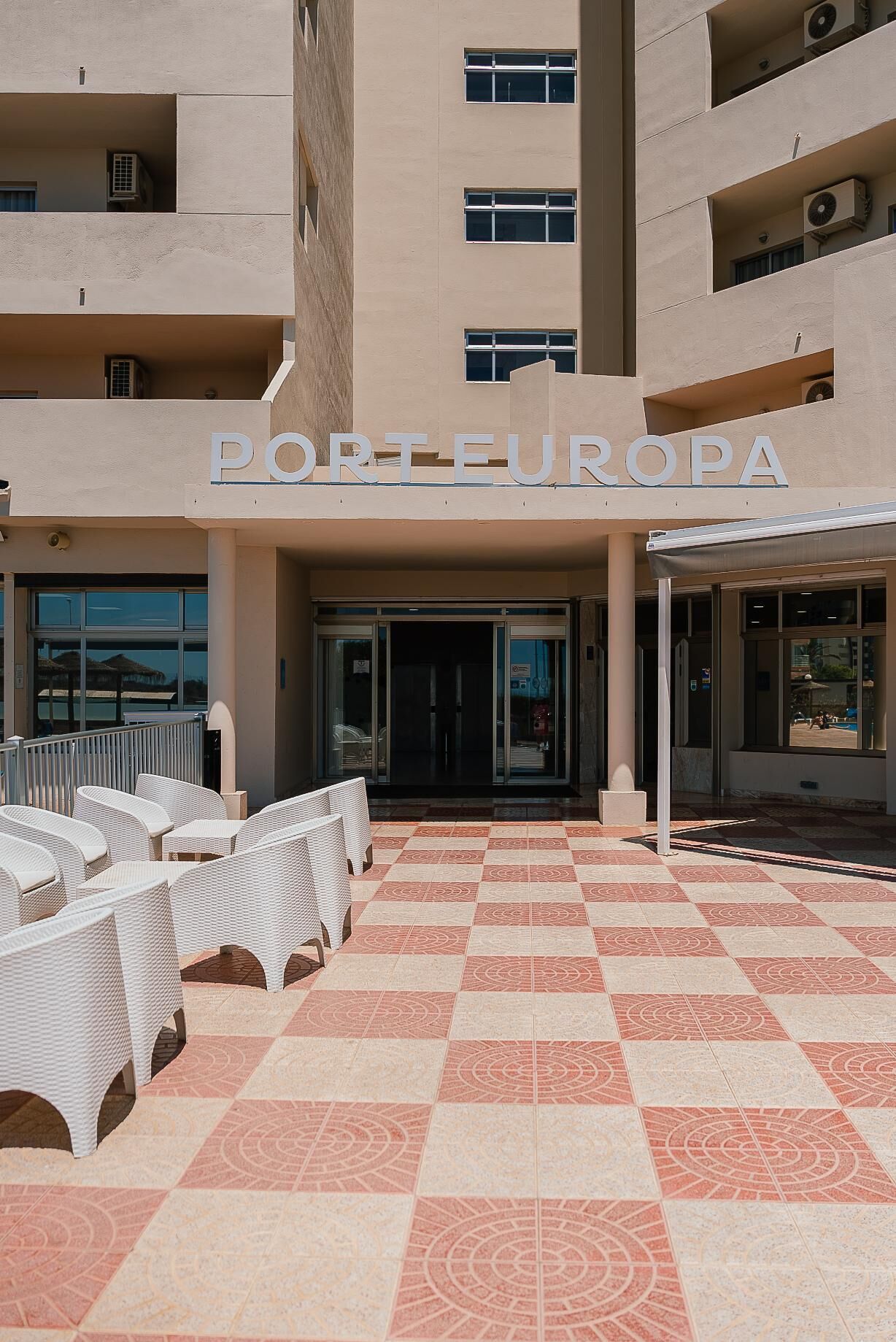 A hotel entrance with a sign reading "PORTEUROPA," outdoor seating with white wicker furniture, and a patterned tile floor.