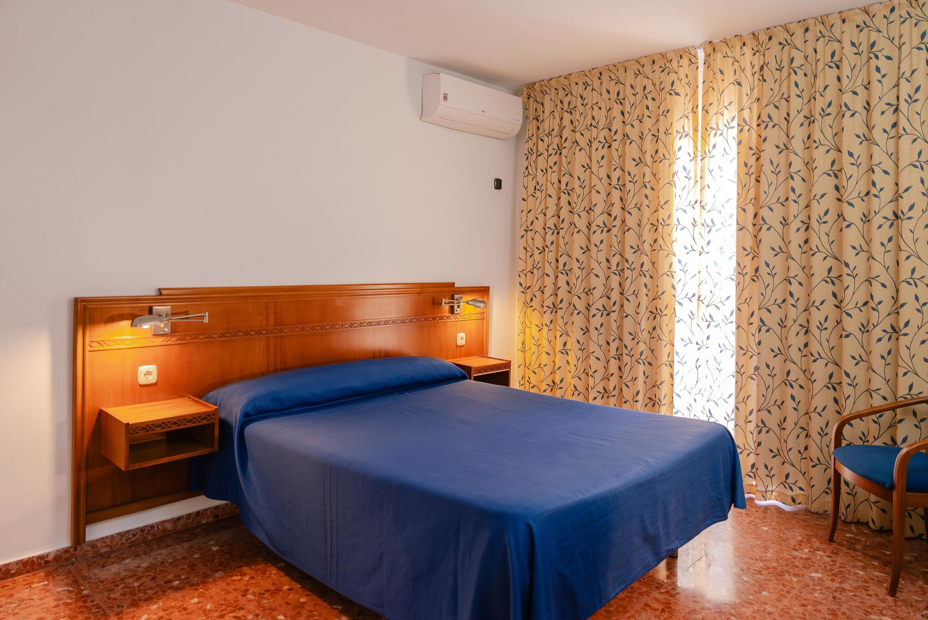 A hotel room with a wooden headboard, a single bed with blue bedding, a nightstand with a lamp, a chair, and patterned curtains.