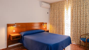 Habitación de hotel con cabecera de madera, una cama individual con ropa de cama azul, una mesita de noche con lámpara, una silla y cortinas estampadas.