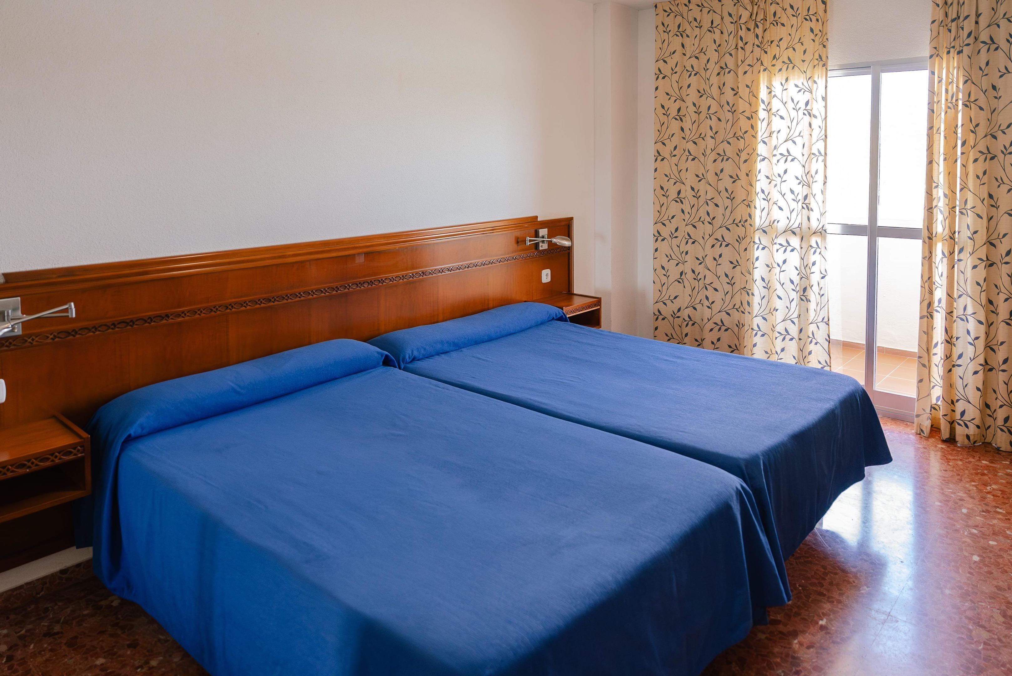 A hotel room with two single beds, a wooden headboard, a nightstand, and a window with curtains.