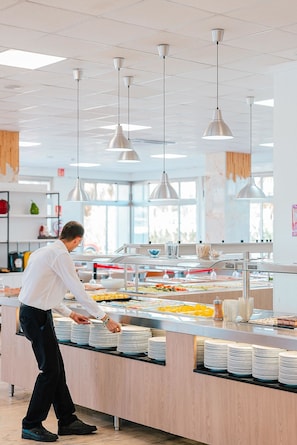 Zona de buffet con varias estaciones de comida, una persona sirviendo alimentos y lámparas colgantes.