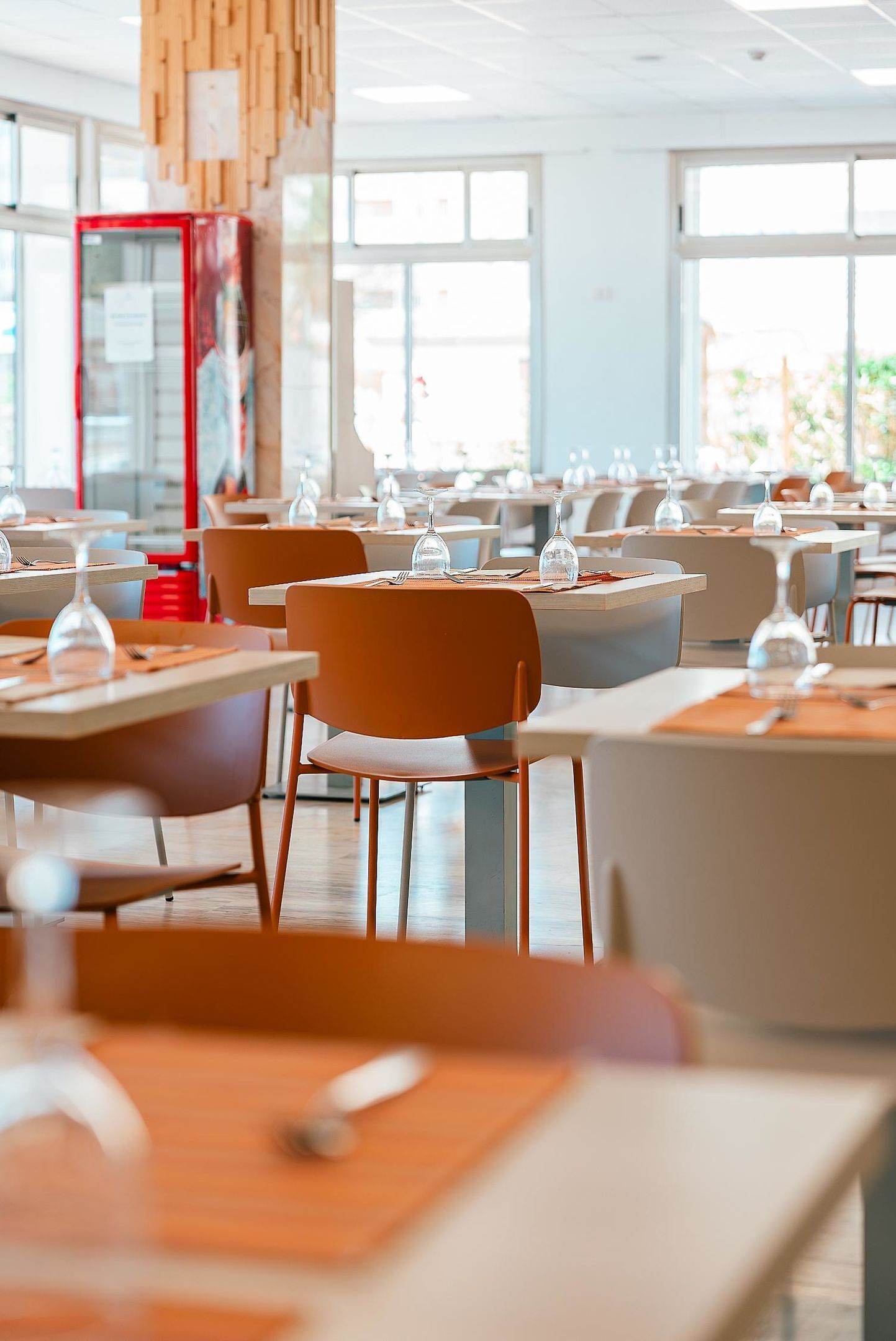 Un comedor moderno con mesas, sillas, ventanas amplias y un armario rojo.