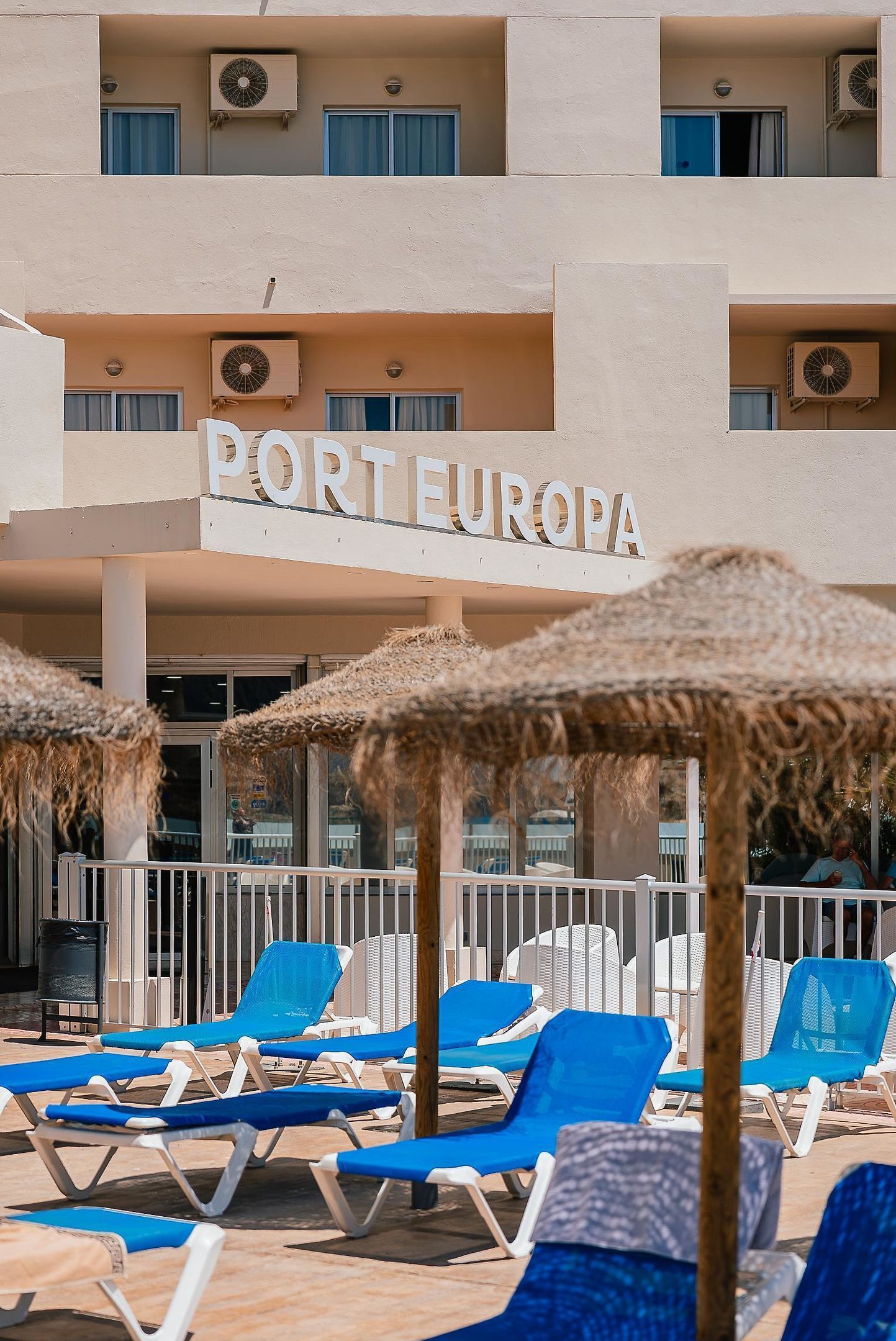 Een hotel genaamd "PORTEUROPA" met meerdere balkons, airconditioning en een zwembadgedeelte met blauwe ligbedden en strooien parasols.