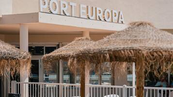 Een hotel genaamd "PORTEUROPA" met meerdere balkons, airconditioning en een zwembadgedeelte met blauwe ligbedden en strooien parasols.